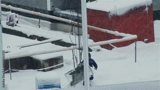Worker in blue winter suit pushing large heavy object through deep snow on outdoor industrial site. Winter storm with falling snow creates challenging conditions for manual labor.