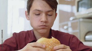 A 15 year old teen bites a sandwich. A young guy eats a sandwich at a table.