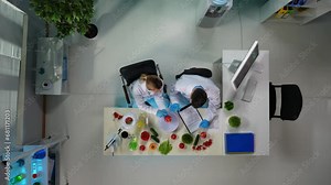 The frame above shows scientists sitting in a laboratory. A woman is doing research, testing, taking samples of an apple and vegetables that are near her. A man sits next to her and helps her