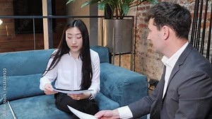 European Asian business partners discussing a commercial proposal in the meeting room. Multinational colleagues sitting on sofa holding contract documents in hands talking negotiating business deal.