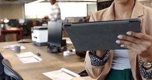 Biracial woman using tablet in modern office, open workspace with colleagues working. Business start up casual entrepreneur unaltered, colleagues collaborating in background, slow motion