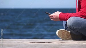 Young woman hands using mobile smart phone at the edge of wooden ocean pier, close up, folded legs, sending sms text messages and browsing internet.