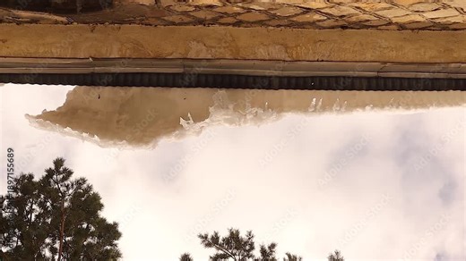 Falling ice and melting icicles from a roof edge with dripping water, highlighting spring thaw, climate change, seasonal danger and urban safety risks. For weather, environment and hazard concepts.