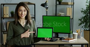 Beautiful woman standing in the office with a tablet device in hands and pointing with a finger on its green screen. Computer screen behind. Chroma key. Indoors