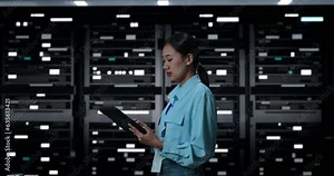 Side View Of Asian Woman Working With Digital Tablet In Server Room Database. Looking Around Inspecting Operating Database Server Rack In Dark Digital High Tech Room