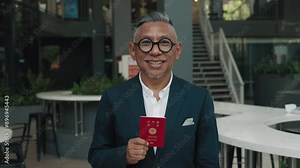 Asian mature man with broad smile holds Japan passport. Smiling male demonstrates passport in vibrant red cover. Senior male dressed in sharp blazer poses for photograph with document ready to travel