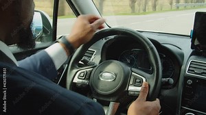 Close-up of black male hands on steering wheel in modern car controlling vehicle, switching on passing beam headlights while man driving car on highway during road trip.