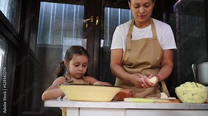 Charming African American woman in chef's apron admiring her cute child, a baby girl who is learning to cook dumplings or vareniki in a summer village kitchen. Mother and daughter baking in kitchen