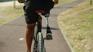 Back view of man with disability riding bicycle in public park. Medium shot of man with disability doing physical activity on sunny day in nature. Disability, sport concept
