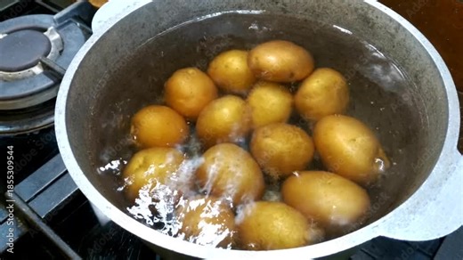 Whole potatoes with their skins are boiled in a metal pan of boiling water. Steam rises from the surface, a cooking process. Boiled potatoes are an ingredient.