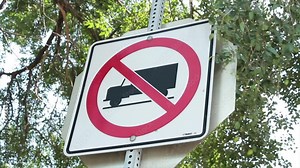 no trucks allowed square sign on metal post with stop sign behind and tree branches blowing behind and in front with sky