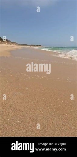 A low-angle vertical view of a pristine coastline with turquoise waves crashing on golden sand, dunes, and blue sky. The wild and famous beach is called Bolonia, in Cadiz, Andalusia, Spain Stock Video Footage - Alamy