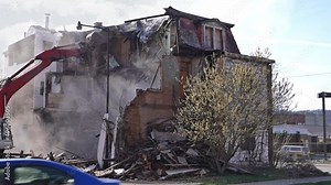 A wide view of a building demolition excavator destroying an old apartment building in a small town. Shot at 60fps for optional slow motion use. With audio.