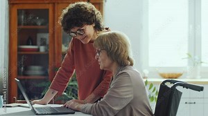 Woman pointing at laptop screen and speaking with senior grandma in wheelchair as teaching her how to use the Internet at home