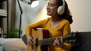 Young woman wears music headphones playing a musical instrument while practicing acoustic guitar and practicing singing sitting on a comfortable sofa in the living room at home.