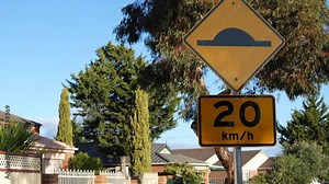 A traffic warning sign indicates the presence of a speed hump on the road with a 20km speed restriction applied in Australia, with a blurry view of suburban houses in the neighborhood.