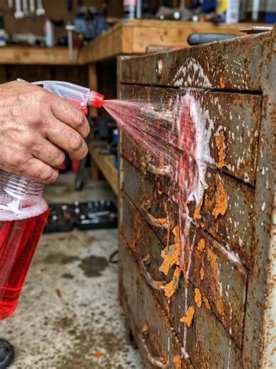 An old, heavily rusted metal cabinet stands in a realistic setting. One realistic adult human hand enters from the left side of the frame holding a fully visible clear plastic spray bottle filled with purple cleaning liquid. As the trigger is pressed, the spray stream stays physically connected to the nozzle and spreads purple foam evenly across 100% of the visible metal surface of the cabinet. Rust visibly flakes off, small rust particles fall downward, and bare iron is gradually revealed benea