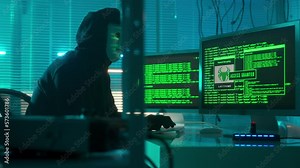 Hacker in a white mask is typing on a computer keyboard and cracking a password. Access denied, access granted. A man in a black hoodie and gloves. The mask reflects green symbols of program codes.