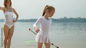 mother and daughter in white bathing suits dancing with gymnastic ribbon on a sandy beach. Summer, dawn