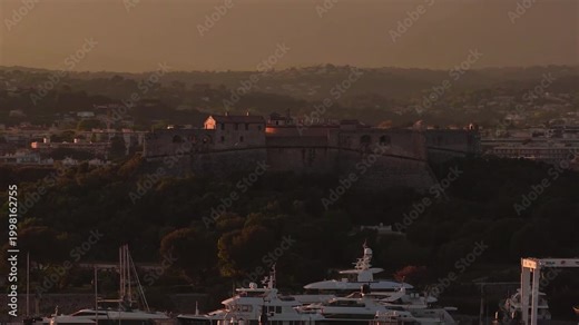 Aerial drone pan sweeps Antibes, France at golden hour. Fort Carre crowns a wooded hill above Port Vauban as superyachts rest in the marina, warm light casts long shadows.