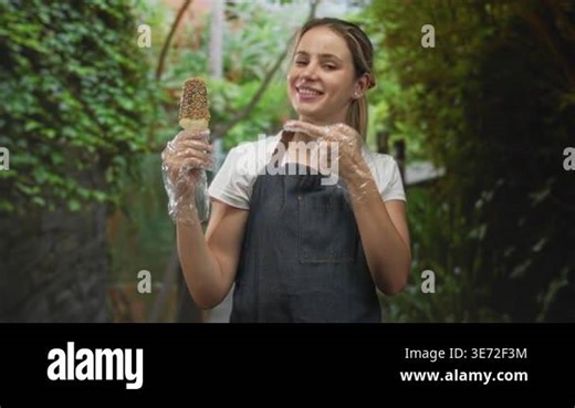 Young blonde woman wearing apron and disposable gloves holding ice cream cone and points finger to treat in park walkway; simple joy Stock Video Footage - Alamy