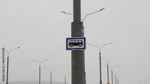 Bus stop sign on pole with falling snow, surrounded by numerous street lamps in muted, snowy landscape. Essence of winter in urban setting, highlighting impact of snow on public transportation.
