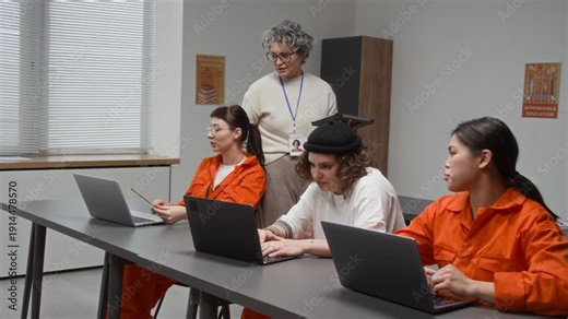 Group of young women as female inmates sitting in row and using laptops with grey haired mature woman overseeing work and assisting during computer programming class in prison education program