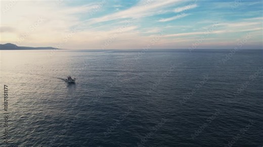 Drone aerial 4k resolution footage of fishing boat sailing away on calm ocean water with a mountain range on the horizon under a soft sunset or sunrise sky. Akamas Paphos Cyprus