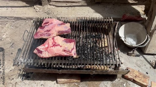 Argentine 'asado de obra' on an improvised metal grill. Raw meat cooking over wood and charcoal fire at a dusty construction site with heavy smoke
