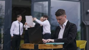 Serious busy young employee in suit sitting on office terrace messaging online on laptop with blurred coworkers talking at background. Portrait of confident concentrated Caucasian man typing e-mail