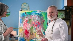 portrait of an old talented man together with his wife paint a modern picture with paints and brush on canvas