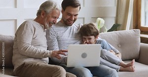 Handsome young man web surfing information, shopping, watching movie on computer with mature older father and little school aged kid son, spending free leisure time with electronic device at home.