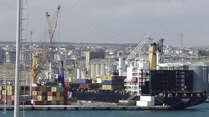 Container ship loading at dock. Crane loads cargo into docked ship at international trade port. Loader machine on pier hauls containers for export onto transport boat at harbor.