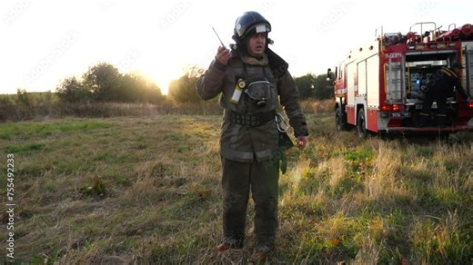 Professional fireman in full equipment talking on walkie-talkie on place at fire near a big red truck. Male firefighter speaking on radio communication against background of fire engine at countryside
