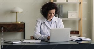 Pensive aged woman doctor sit at table do paperwork at clinic office make scientific research using laptop pc paper history sheets. Mature female medical agent work with insurance policies of patients