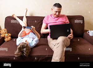Happy young father sit on computer using laptop with son having a work video call. Smiling dad and little boy child legs up at home rest on sofa busy Stock Photo - Alamy