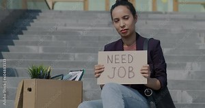 Slow motion of unemployed young woman holding Need Job sign sitting on stairs of office building in city. Unemployment and crisis center.