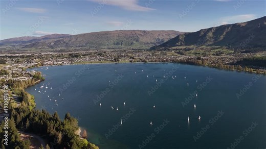 A cinematic 4K 60fps backward drone shot revealing a fleet of sailboats during a regatta on the blue waters of Lake Wanaka, with the Southern Alps in the distance, New Zealand