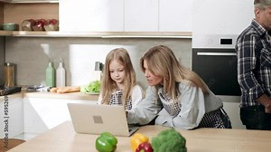 Smiling blond mother help her cute little daughter with homework on laptop computer at distance remote education while busy father has working business call by cellphone at domestic kitchen at home
