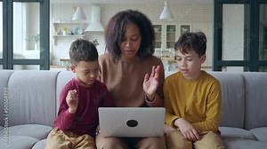 Happy family of three bonding using laptop, relaxing on the couch, greeting, waving hands during a video call via application. Single mother sitting between adorable children having fun at the laptop