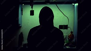 Silhouette of a mysterious hacker wearing a hoodie playing his computer and his phone full of green programming codes in a dark room