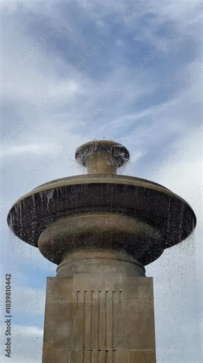 The top section of the stone fountain is visible with water cascading over its rounded edge. The background shows a cloudy sky, contrasting with the stone texture of the fountain.
