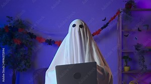 Funny halloween ghost in white sheet with black eyes in dark room working on laptop, typing text surfing on the web. Happy halloween spooky concept.
