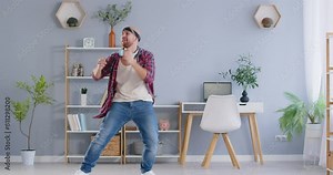 Young man having fun after completing work. Happy man in a casual shirt, jeans and bandana headband, sitting at a desk at home, finishes work on his laptop computer, stands up and starts dancing