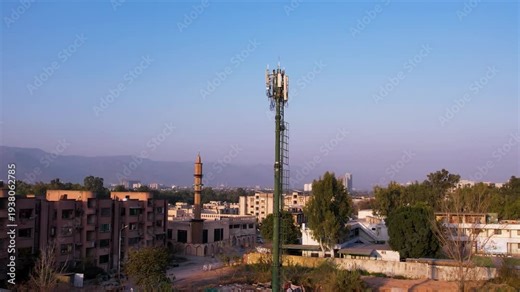 A tall cell tower stands among buildings and trees in a sunlit urban area