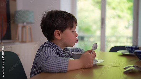 Young boy sitting at the dining table, lost in thought while holding a spoon and bowl, creating a peaceful and contemplative moment during a quiet meal at home