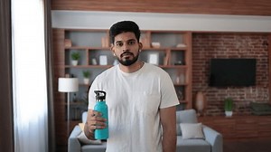 Tired indian man in sport clothes wiping sweat and drinking water during morning exercise. Active young guy having regular workout during free time at home.