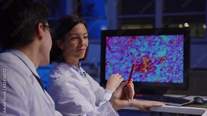 Zoom in of young male scientist making notes and examining test tube with red liquid with female colleague showing him magnified microorganisms on computer screen in biochemistry laboratory