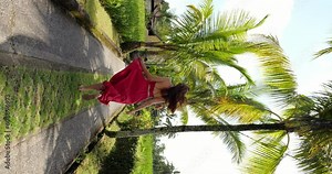 Wonderful vacation life, happy tourist woman run at tropical alley, red dress skirt fly in air, slow motion vertical shot. Camera follow behind lady, she glance back once and turn around at end
