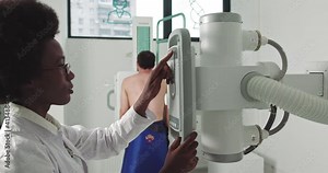 Female Technician adjusts X-Ray Machine in Medical Cabinet. Scanning for Fractures and Broken Limbs. In the Hospital, Man Standing Face Against the Wall While Medical Technician Adjusts X-Ray Machine